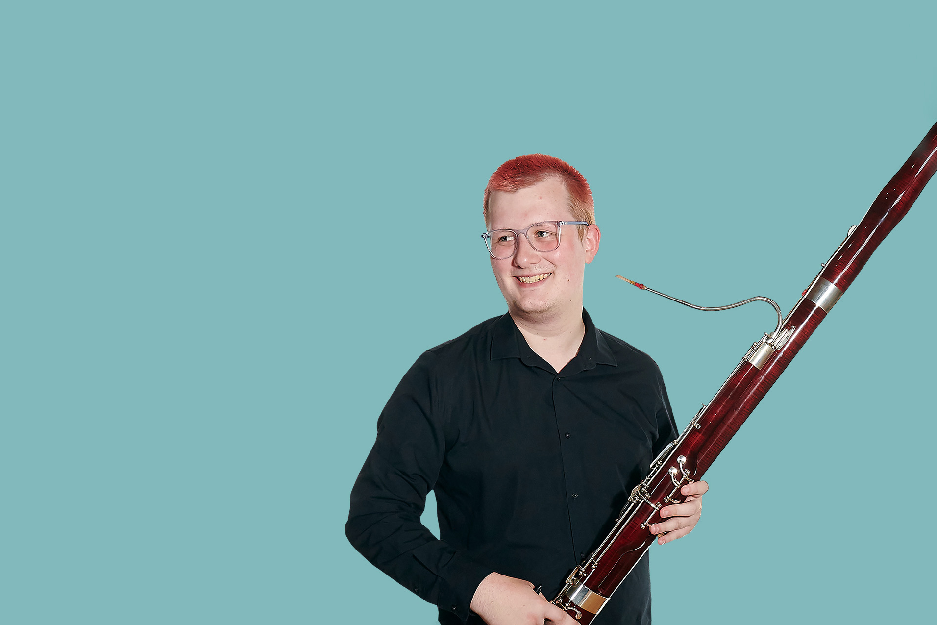 A male student wearing a black shirt, smiling, holding a bassoon.